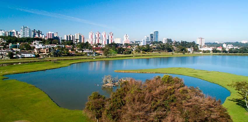 Vista de cima do Parque Barigui, em que é apresentado o lago que o compõe, as áreas verdes e os prédios do entorno